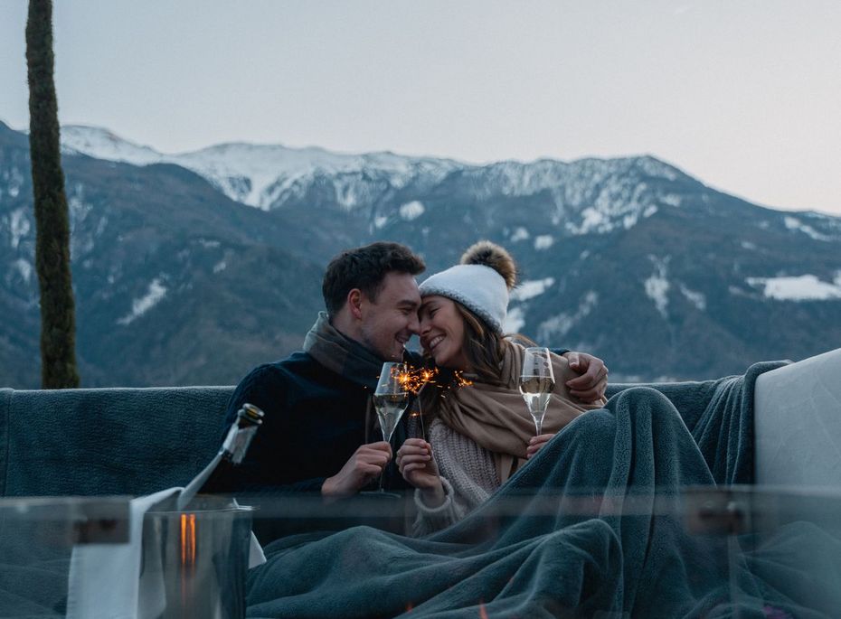 Paar am Rooftop-Spa mit Feuerstelle Ein Paar sitzt eingekuschelt am Rooftop-Spa vor einer Feuerstelle mit Bergblick im Hintergrund.