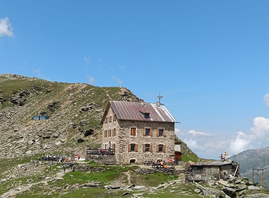 Berghütte auf felsiger Alm im Naturns-Blog Eine Berghütte steht auf einer felsigen Alm unter klarem Himmel, mit Wanderern auf dem Weg und weitem Blick in die Berge.
