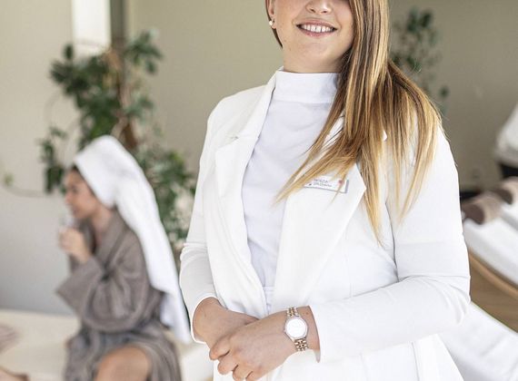 A smiling staff member in a white coat stands in a relaxation room with guests resting in the background.