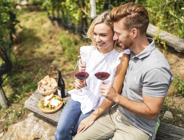 Ein Paar sitzt auf einer Holzbank und hält zwei Gläser Rotwein beim Picknick im Grünen.