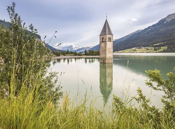 Una torre in pietra si riflette in un lago alpino calmo, con canneti, pendii verdi e cime innevate sullo sfondo.