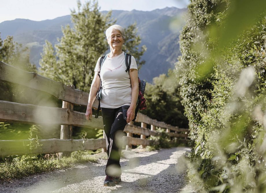 An older woman walks along a gravel hiking path with mountains in the background and greenery in the foreground.
