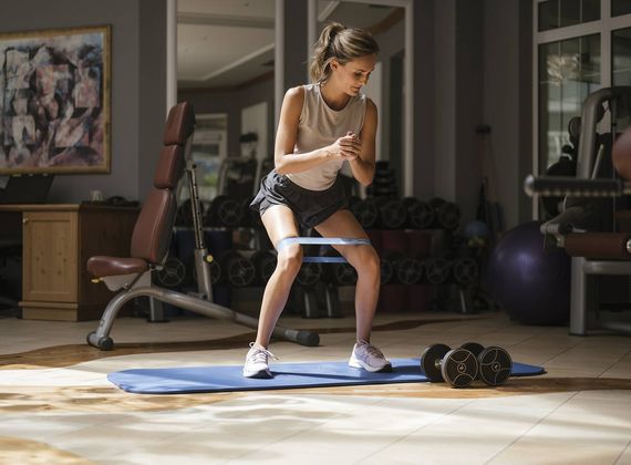 A woman performs a resistance band squat on a blue mat in the gym.