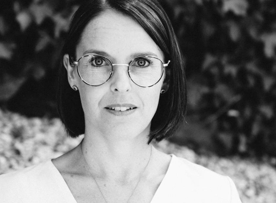 Porträt einer Frau mit Brille im Preidlhof Blog Eine Frau mit runder Brille blickt frontal in die Kamera, vor einem unscharfen Hintergrund.