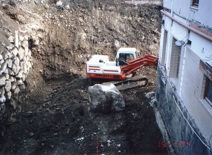 Ein Bagger arbeitet in einer tiefen Baugrube neben einem Gebäude und bewegt einen großen Stein.