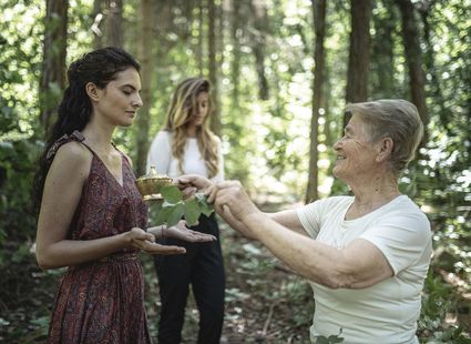 Eine Frau reicht einer anderen im Wald mit einer Schale und Blättern ein kleines Ritualobjekt.