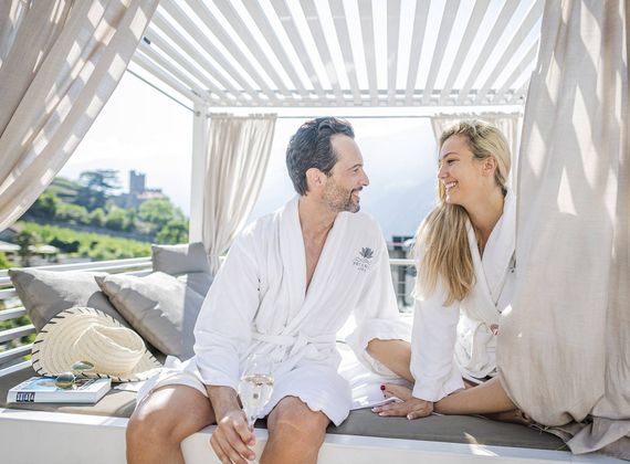 Ein Paar im Bademantel sitzt lächelnd auf einer Daybed-Liege unter einer Pergola.