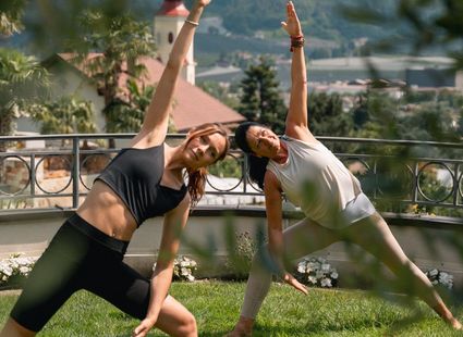 Zwei Frauen machen Yoga auf einer Wiese mit Bergblick und Kirchturm im Hintergrund.