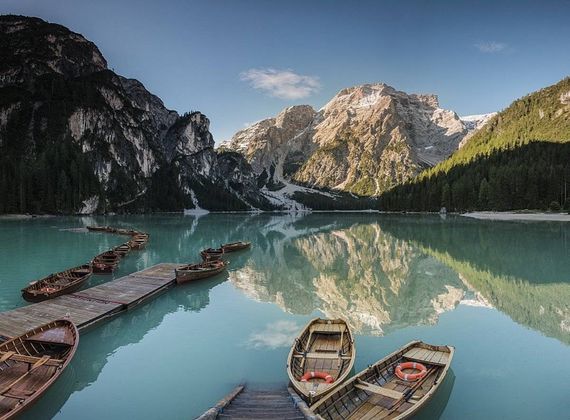 Barche di legno sono ormeggiate a un pontile su un lago di montagna limpido, con pendii boscosi e cime rocciose sullo sfondo.