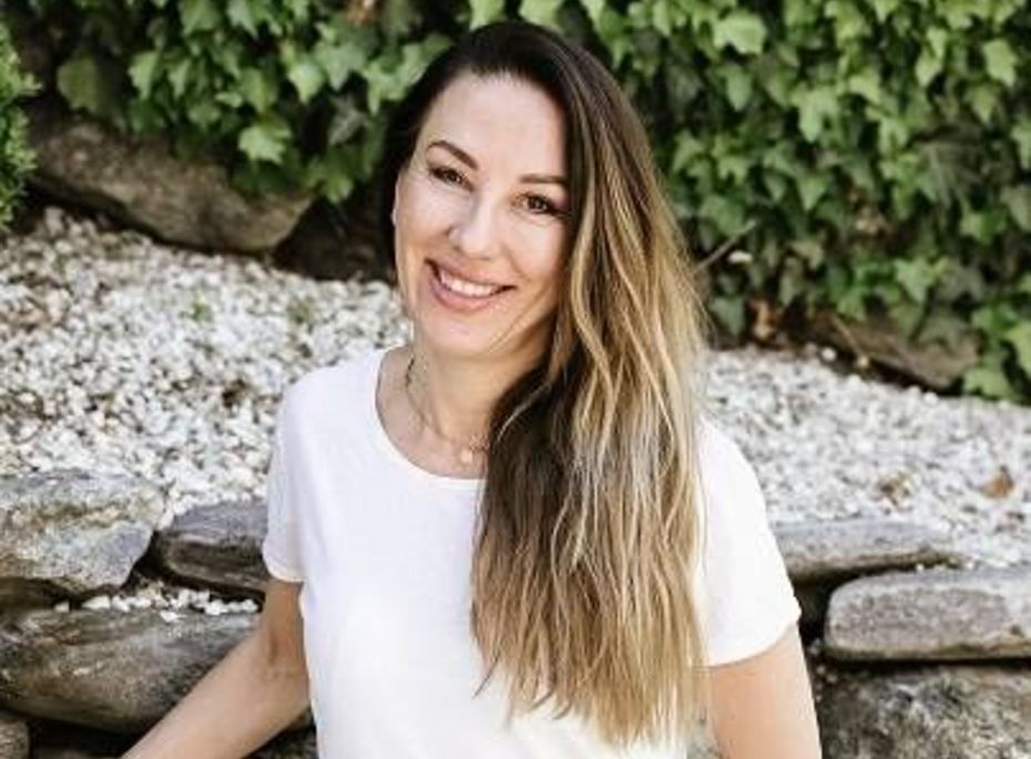 Eine Frau mit langem Haar lächelt vor einer Steinmauer und grünem Blätterhintergrund im Garten.