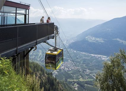 Eine Seilbahn fährt an einer Terrasse mit zwei Personen vorbei, darüber öffnet sich der Blick ins Tal und auf die Berge.
