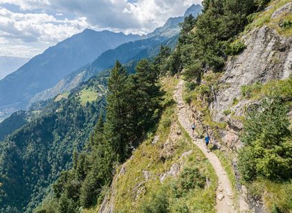 Zwei Wanderer gehen auf einem schmalen Pfad entlang eines steilen Berghangs mit Blick ins Tal.