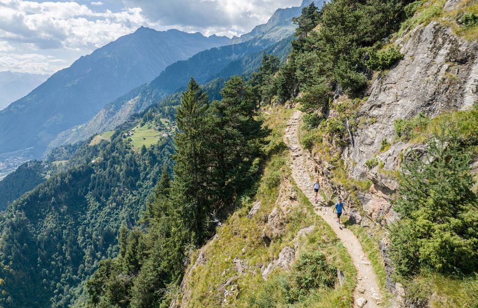 Zwei Wanderer gehen auf einem schmalen Pfad entlang eines steilen Berghangs mit Blick ins Tal.