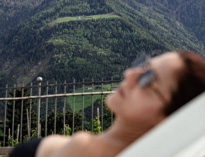 A woman lies blurred in the foreground on a relaxation lounger, with a green mountain slope and fence behind her.