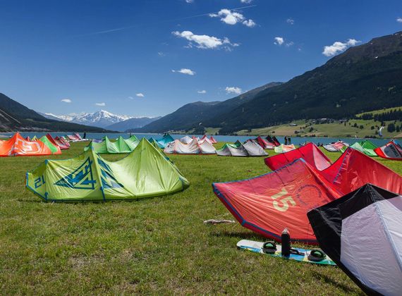Colorful kites and tents are set out on a grassy alpine meadow with mountains in the background.