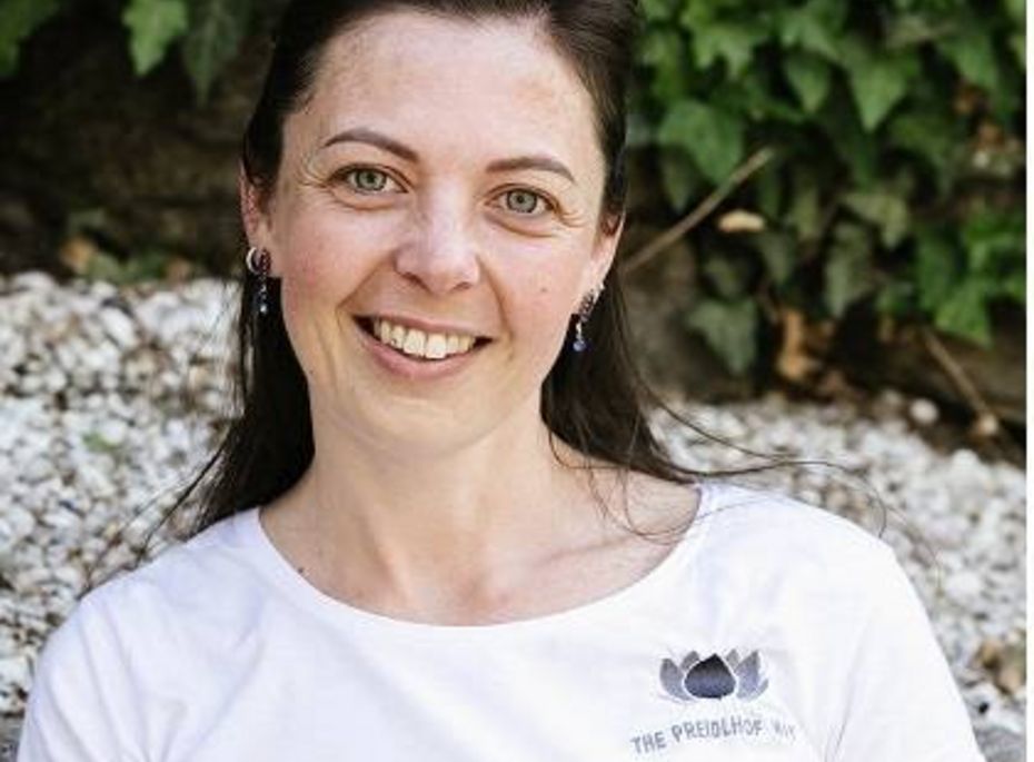 Mitarbeiterin im Preidlhof vor grünem Hintergrund Eine lächelnde Mitarbeiterin mit weißem T-Shirt sitzt vor einer grünen Pflanzenwand.