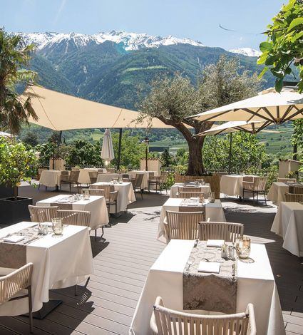Terrazza ristorante all'aperto con tavoli apparecchiati sotto grandi ombrelloni e vista sulle montagne.