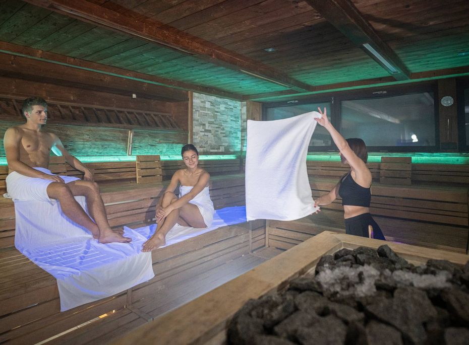 Sauna-Aufguss im Wellnesshotel in Naturns Eine Saunameisterin schwenkt ein Handtuch vor zwei Gästen in einer Holzsauna mit beleuchtetem Ofen.