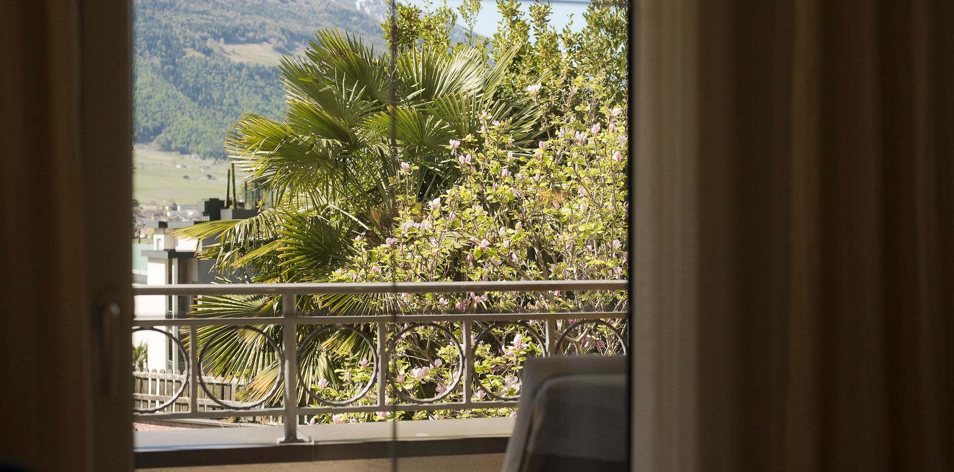 Durch die geöffneten Vorhänge blickt man von der Suite auf den Balkon mit Geländer, Palmen und Berglandschaft.