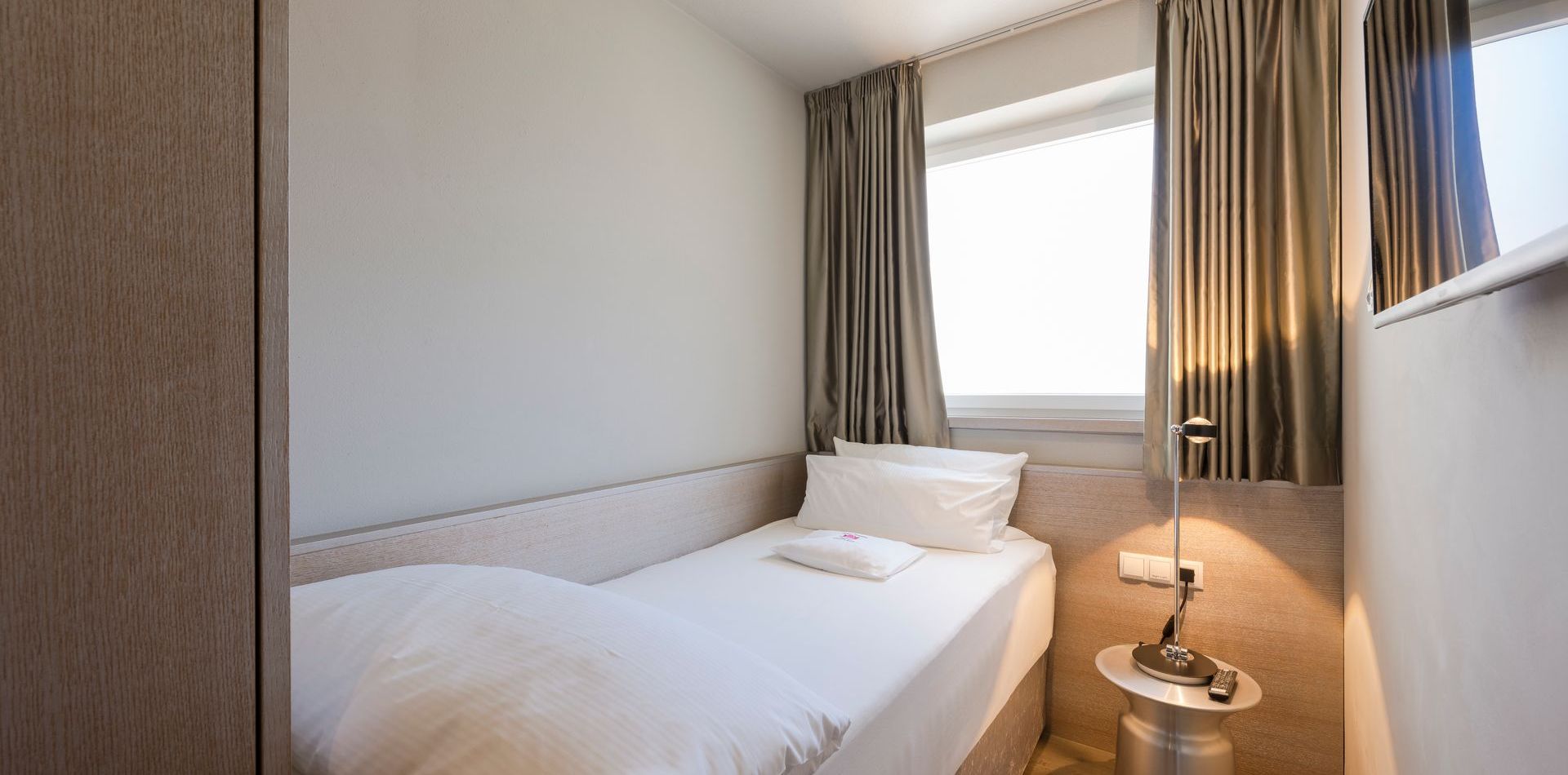 Ein kompaktes Einzelzimmer mit Bett, Fenster, Vorhängen und Nachttisch in der Luxury Suite You & Me Premium.