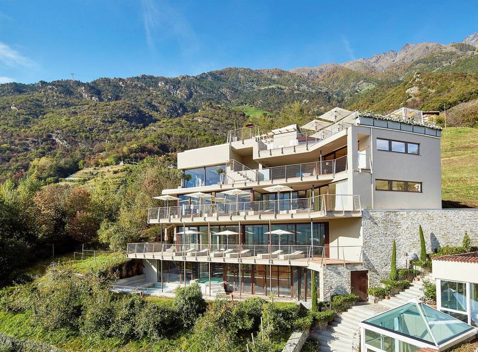 Ein modernes Hotel mit mehreren Terrassen liegt am Hang vor einer grünen Berglandschaft.
