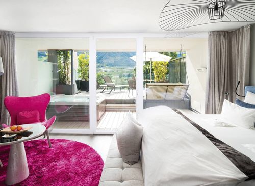 A bright suite with a bed, pink lounge chair, and glass doors opening to a terrace and mountain view.