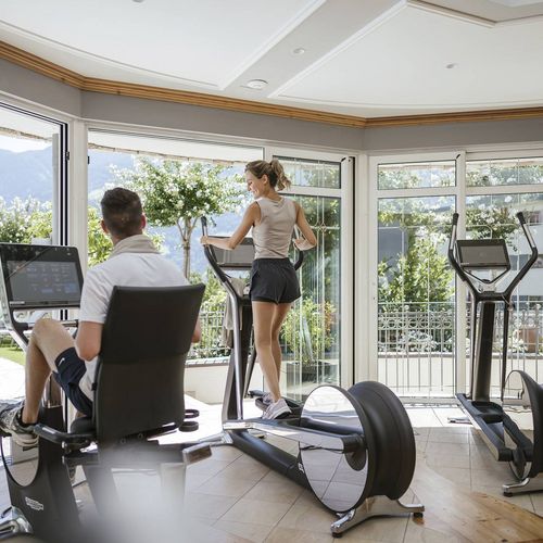 Eine Frau trainiert auf dem Crosstrainer, während ein Mann auf einem weiteren Gerät im hellen Fitnessraum sitzt.