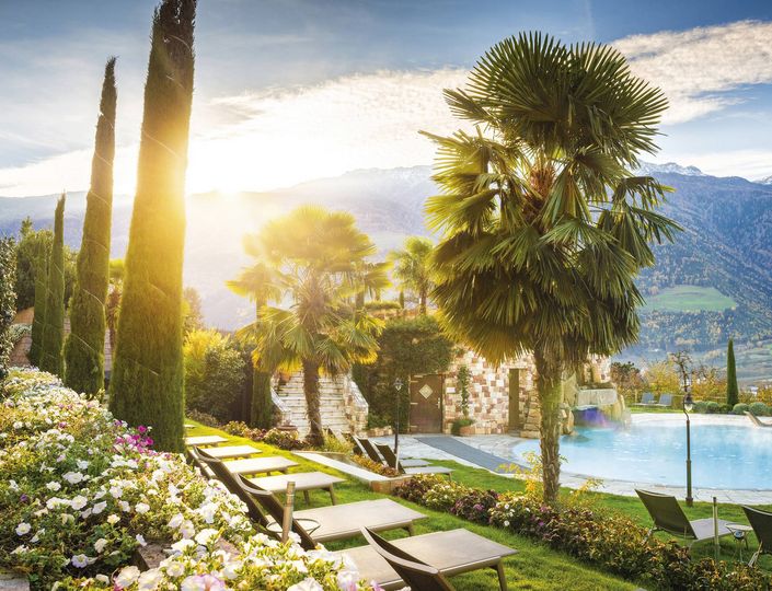 Ein Infinity Pool liegt in einer gepflegten Gartenanlage mit Liegen, Palmen und Bergblick im warmen Abendlicht.