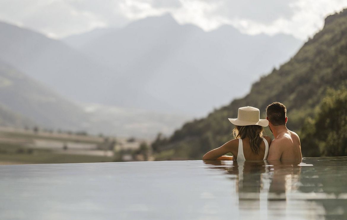 A couple sits in an infinity pool facing a mountain valley view.