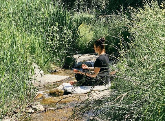 A woman sits on stone slabs by a small stream in a grassy outdoor relaxation area.