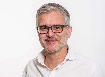 Portrait of a smiling male medical spa team member wearing glasses and a white shirt against a plain background.