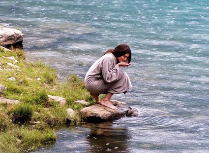 Eine Frau kauert barfuß auf einem Felsen am Ufer eines Sees und blickt ins Wasser.