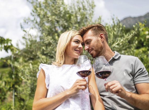 Ein Paar stößt im Garten mit Rotwein an und sitzt entspannt zwischen grünen Bäumen.
