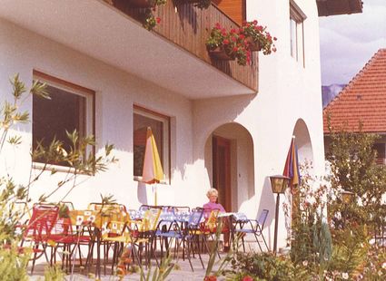 Eine Hotelterrasse mit bunten Stühlen, Sonnenschirmen und einer sitzenden Frau vor dem Gebäude.