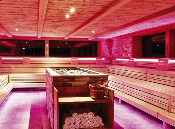 Eine helle Sauna mit Holzsitzecken, zentralem Ofen und aufgerollten Handtüchern im Preidlhof.