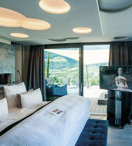 Ein modernes Hotelzimmer mit großem Bett, TV und Balkon mit Blick auf die Berge und das Tal.