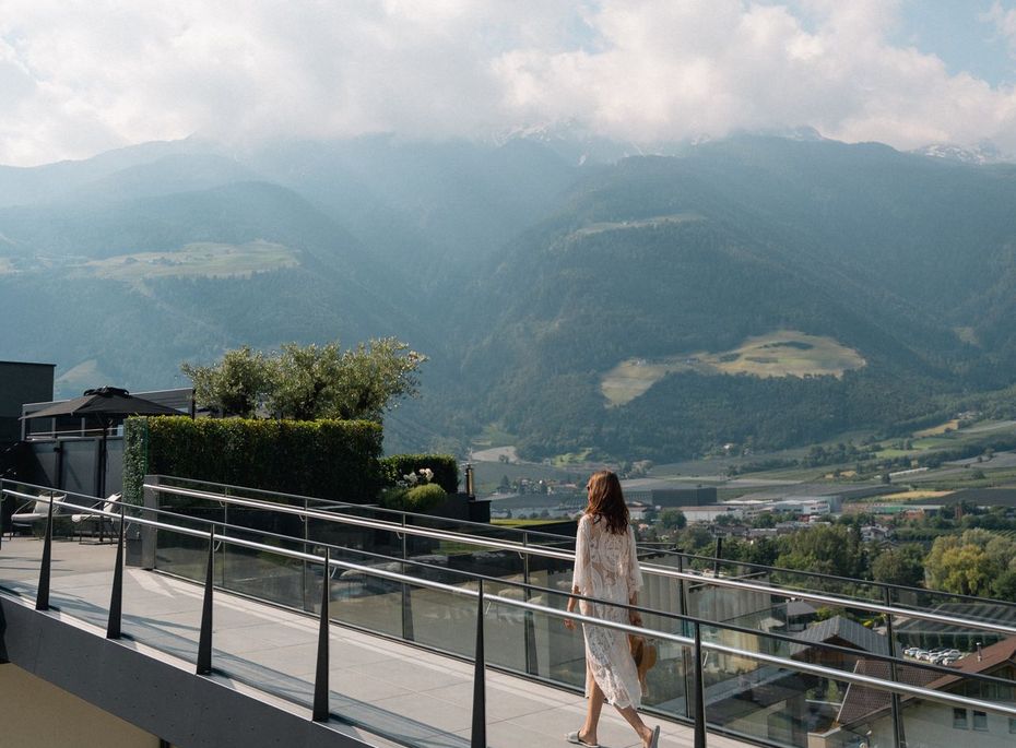 Eine Frau geht über die Dachterrasse mit Glasgeländer und Blick auf das Tal und die Berge.