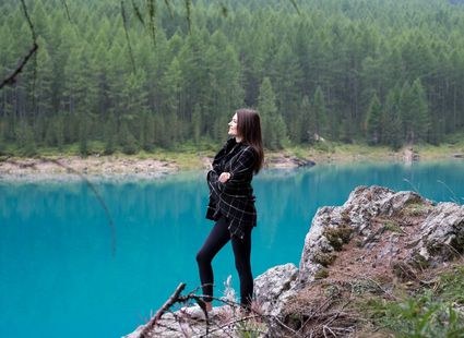 Eine Frau steht auf einem Felsen am türkisblauen Seeufer vor dichtem Nadelwald.