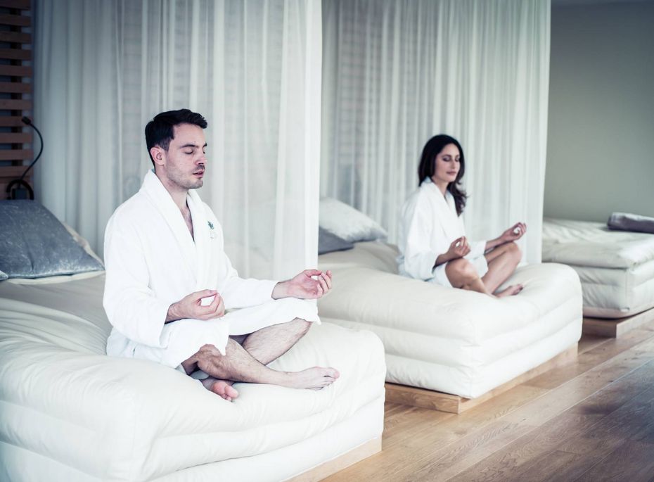 Wellnesshotel: Meditation auf Liegen im Ruheraum Ein Mann und eine Frau sitzen in weißem Bademantel auf Liegen im Ruheraum und meditieren.