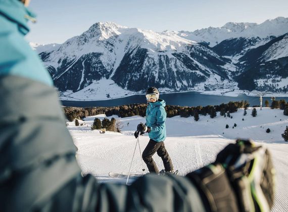 Eine Skifahrerin steht auf einer Piste mit Blick auf die verschneiten Berge und den See im Hintergrund.