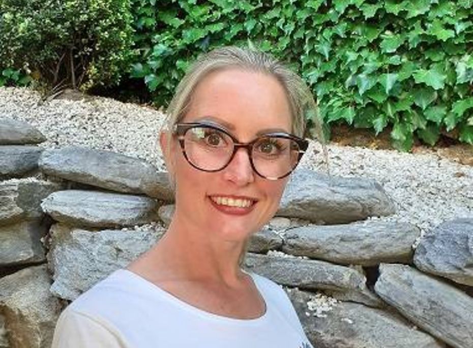 Porträt einer Mitarbeiterin im Preidlhof-Garten Eine lächelnde Mitarbeiterin mit Brille steht vor einer Natursteinmauer und grünem Efeu im Garten.
