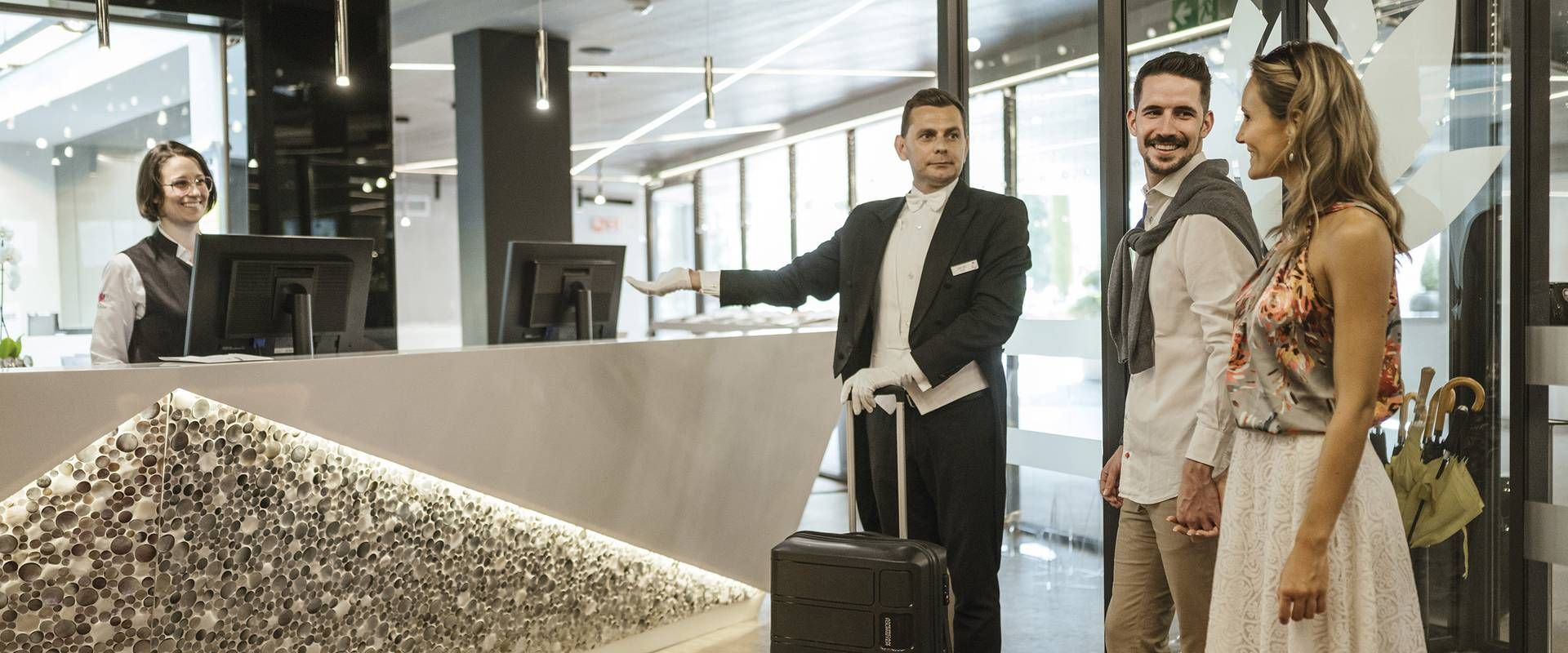 An der Hotelrezeption stehen ein Gepäckträger und zwei ankommende Gäste mit Koffern.