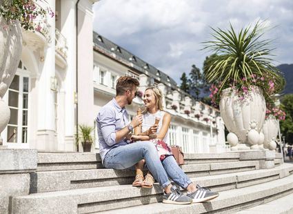 Ein Paar sitzt mit Eis auf den Treppen vor dem Wellnesshotel, daneben stehen große Pflanzgefäße und das Gebäude im Hintergrund.