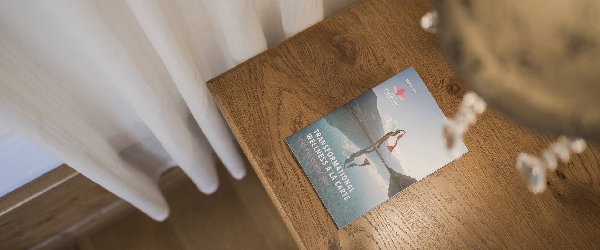 Una brochure del wellness retreat è appoggiata su un tavolo in legno accanto a tende leggere e a un oggetto decorativo sfocato.