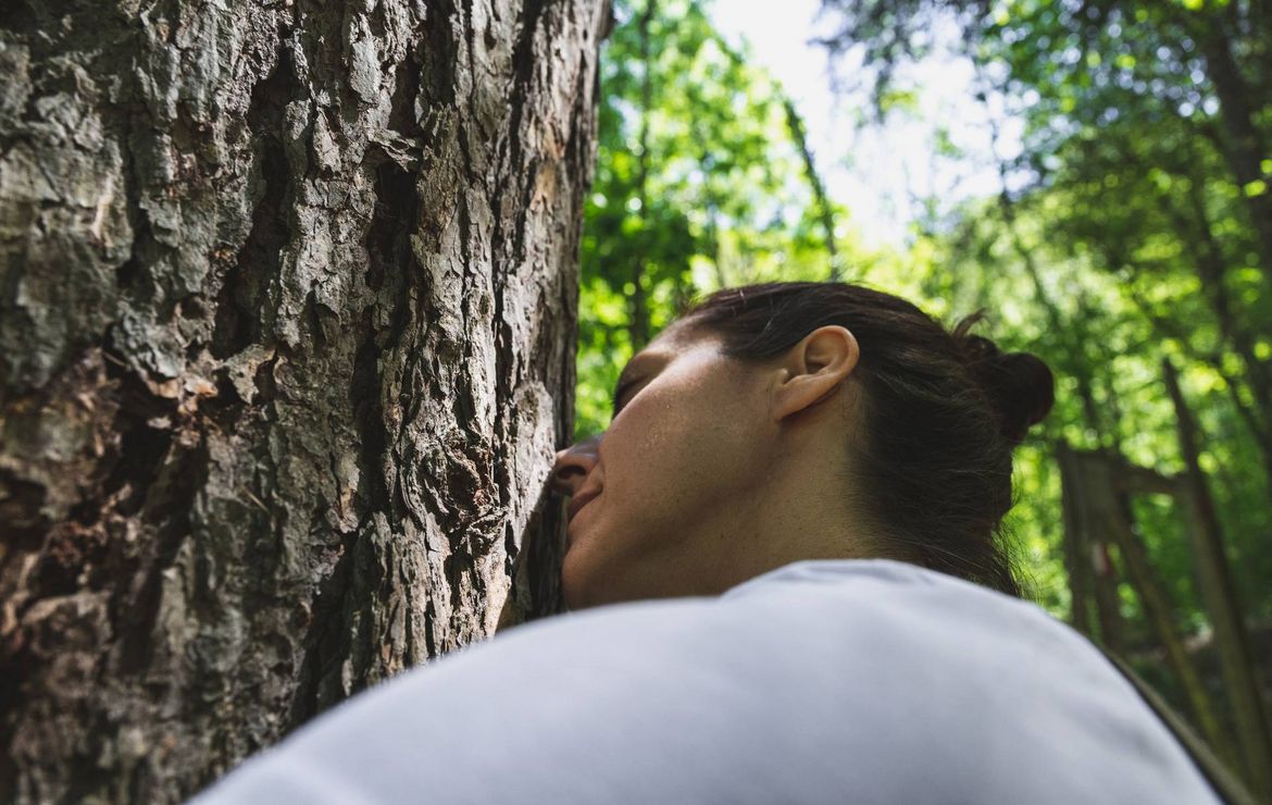 Una donna si appoggia a un albero in un bosco, con la luce del sole tra gli alberi verdi intorno a lei.