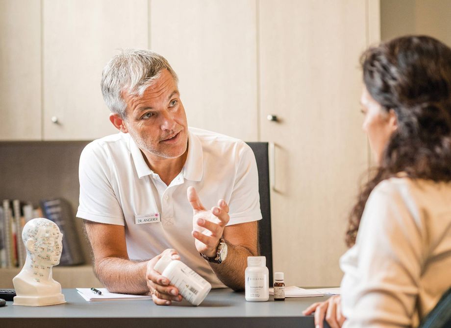 A spa therapist speaks with a guest across a desk, with wellness product bottles and a model head in the room.