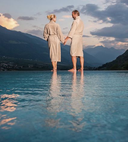 Ein Paar steht im Infinity-Pool und hält sich an den Händen, mit Bergkulisse im Abendlicht im Hintergrund.