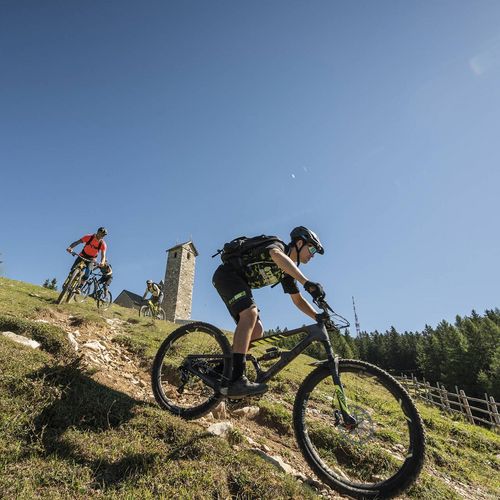 Zwei Mountainbiker fahren auf einem steilen Wiesenweg bergab, im Hintergrund stehen ein kleiner Turm und Wald.