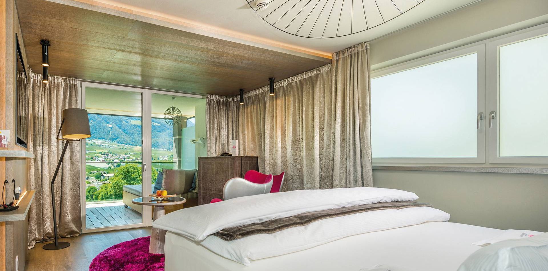 A bright hotel room with twin beds, a balcony door, curtains and a mountain view.