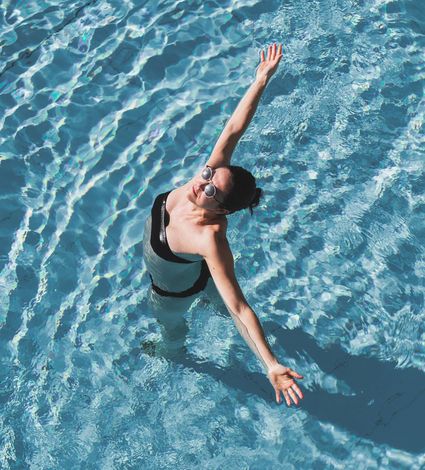 A woman in sunglasses stands in an outdoor pool with her arms raised in the water.